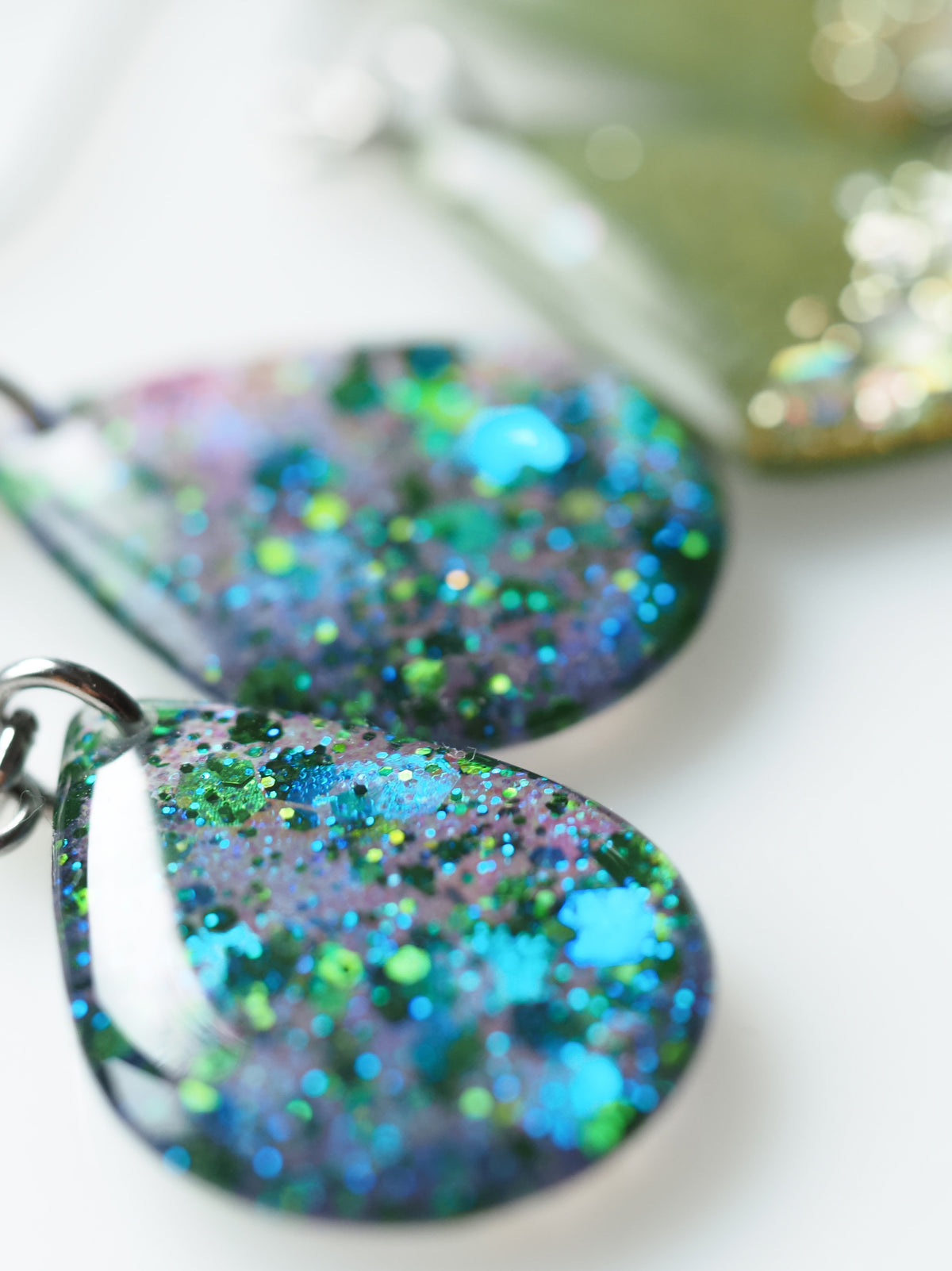 Close up of the Clio drop earrings in iridescent blue and holographic green glitter on a white surface with a pair of green Persephone glitter earrings slightly out of focus in the background

