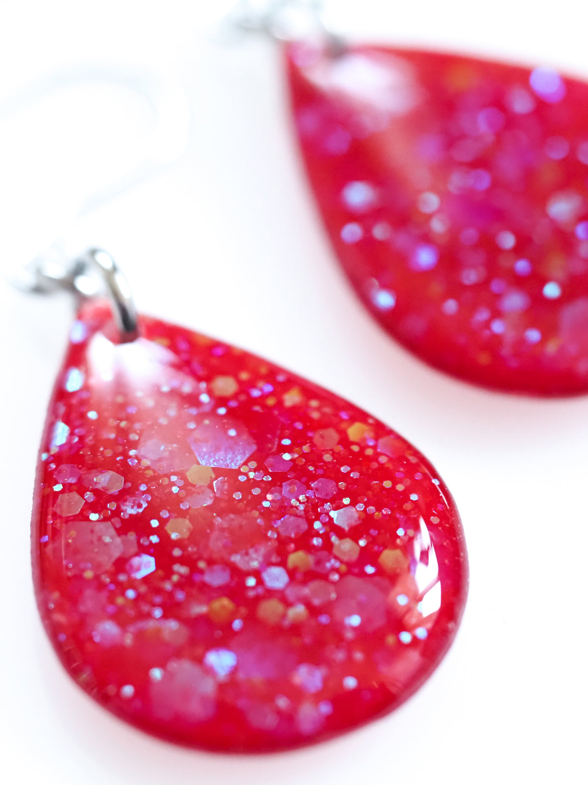 Close up of the Clio drop earrings in red with iridescent colour shifting glitter on a white background