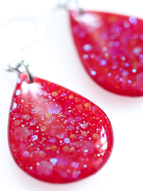Close up of the Clio drop earrings in red with iridescent colour shifting glitter on a white background