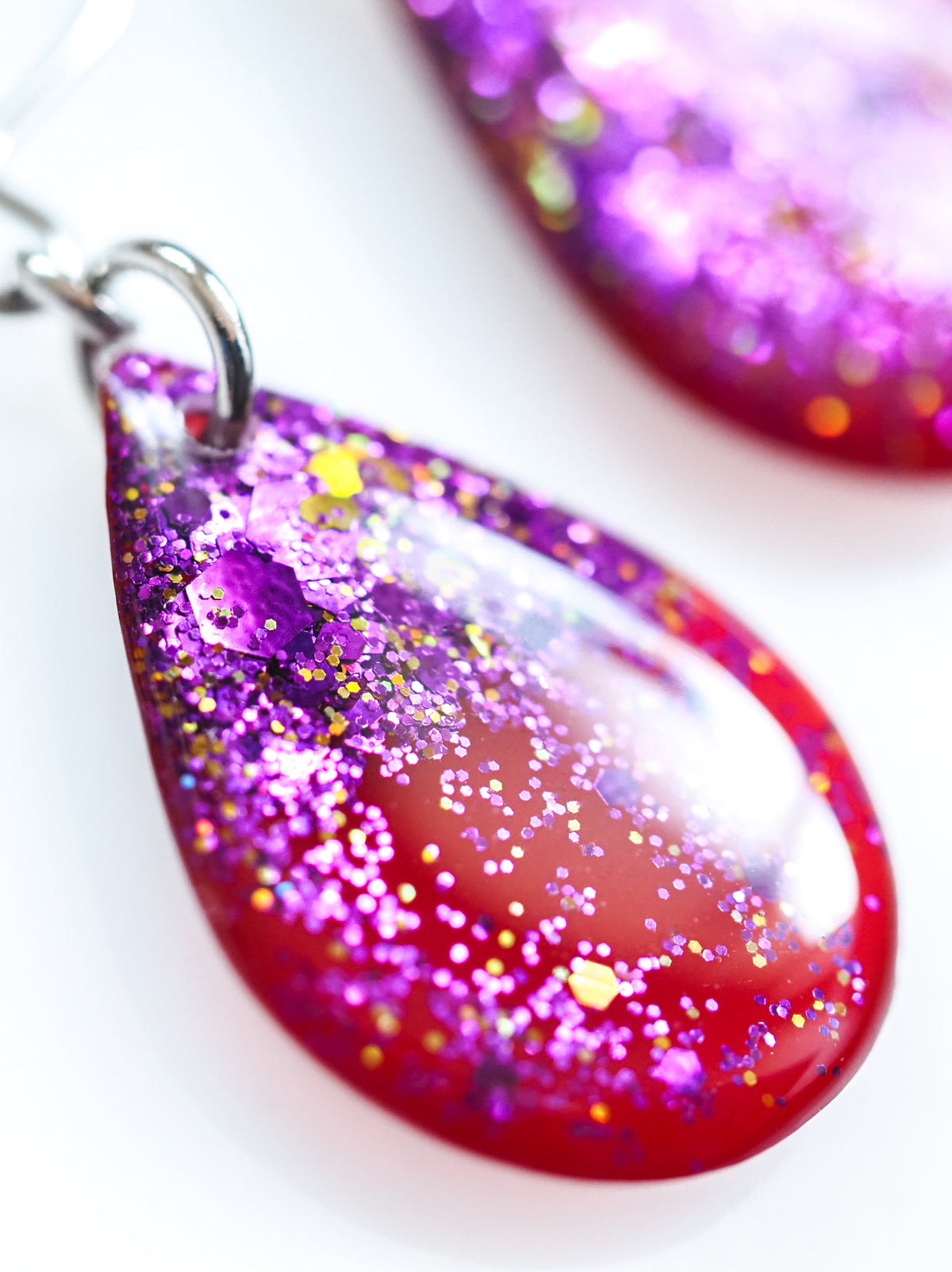 Close up of the Clio drop earrings in red with purple and gold holographic colour shifting glitter on a white background