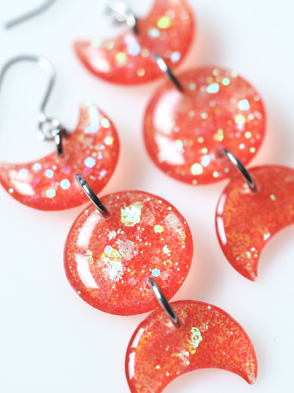 Close up of the Hecate triple moon drop earrings in transparent orange with iridescent glitter on a white background