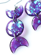Close up of the Hecate triple moon drop earrings in transparent purple with iridescent glitter on a white background