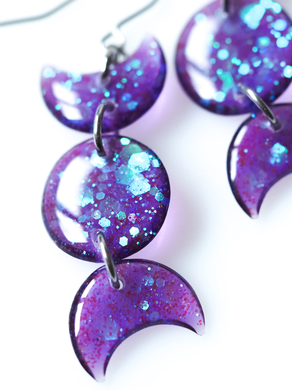 Close up of the Hecate triple moon drop earrings in transparent purple with iridescent glitter on a white background
