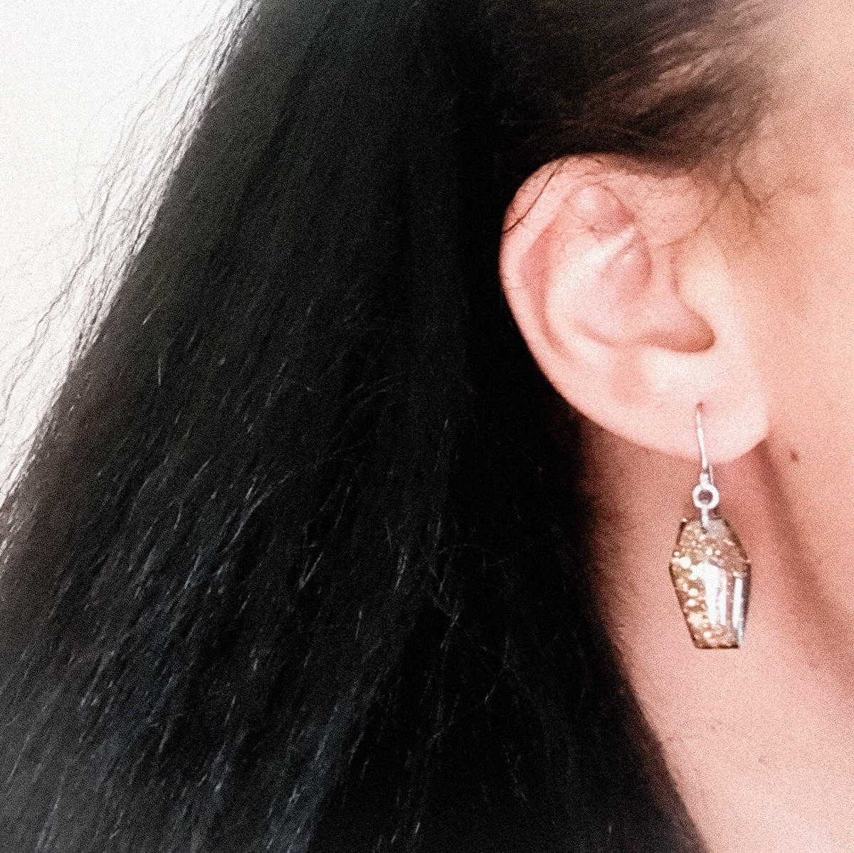 A pair of Morrigan coffin drop earrings, shown being worn. The wearers face is out of shot with hair pulled back to display the earring. 