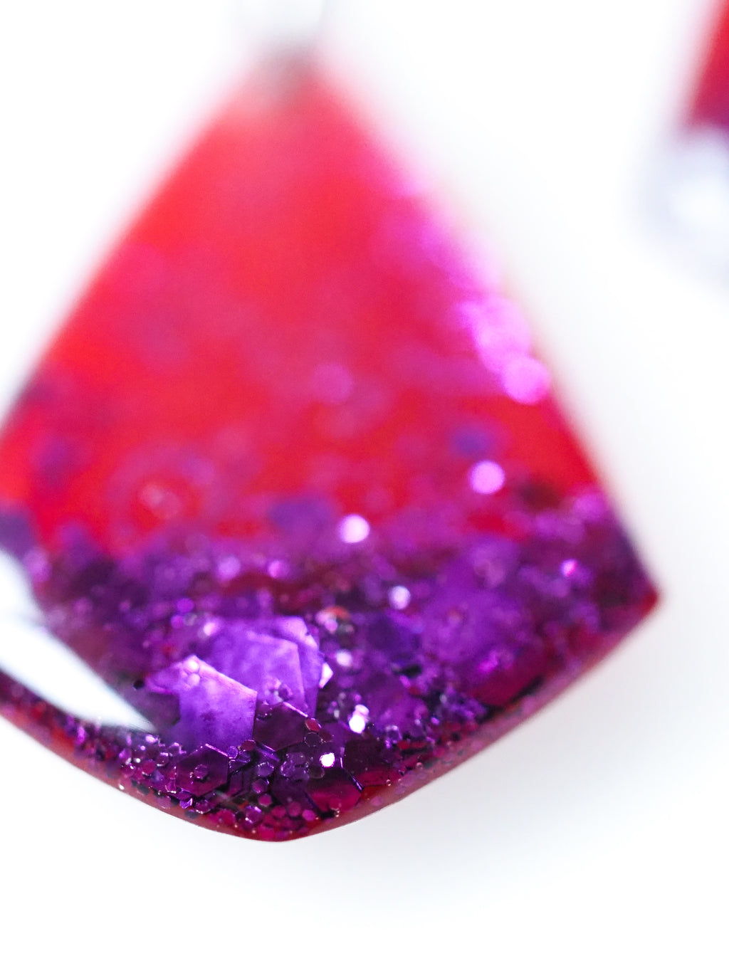 Macro close up of Persephone drop earrings in red with purple metallic glitter on a white background
