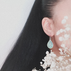 A pair of Clio teardrop shaped drop earrings, shown being worn. The wearers face is out of shot with hair pulled back to display the earring. There are flowers in the foreground.