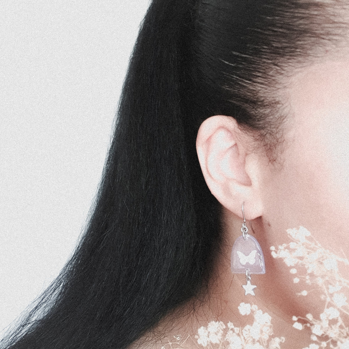 A pair of Nyx drop earrings, shown being worn. The wearers face is out of shot with hair pulled back to display the earring. There are some small flowers in the image foreground framing the earring.
